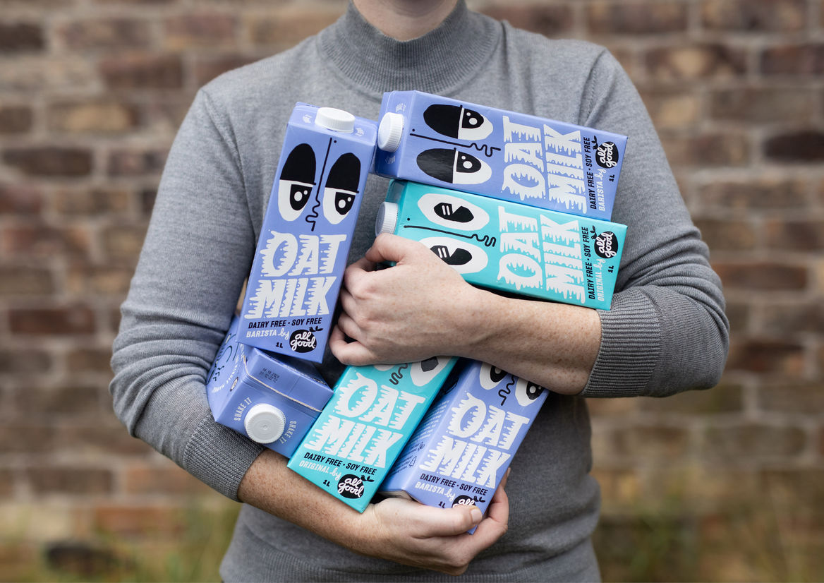 A person in a light grey turtleneck standing in front of a brick wall, holding an armful of cartons of All Good oat milk.