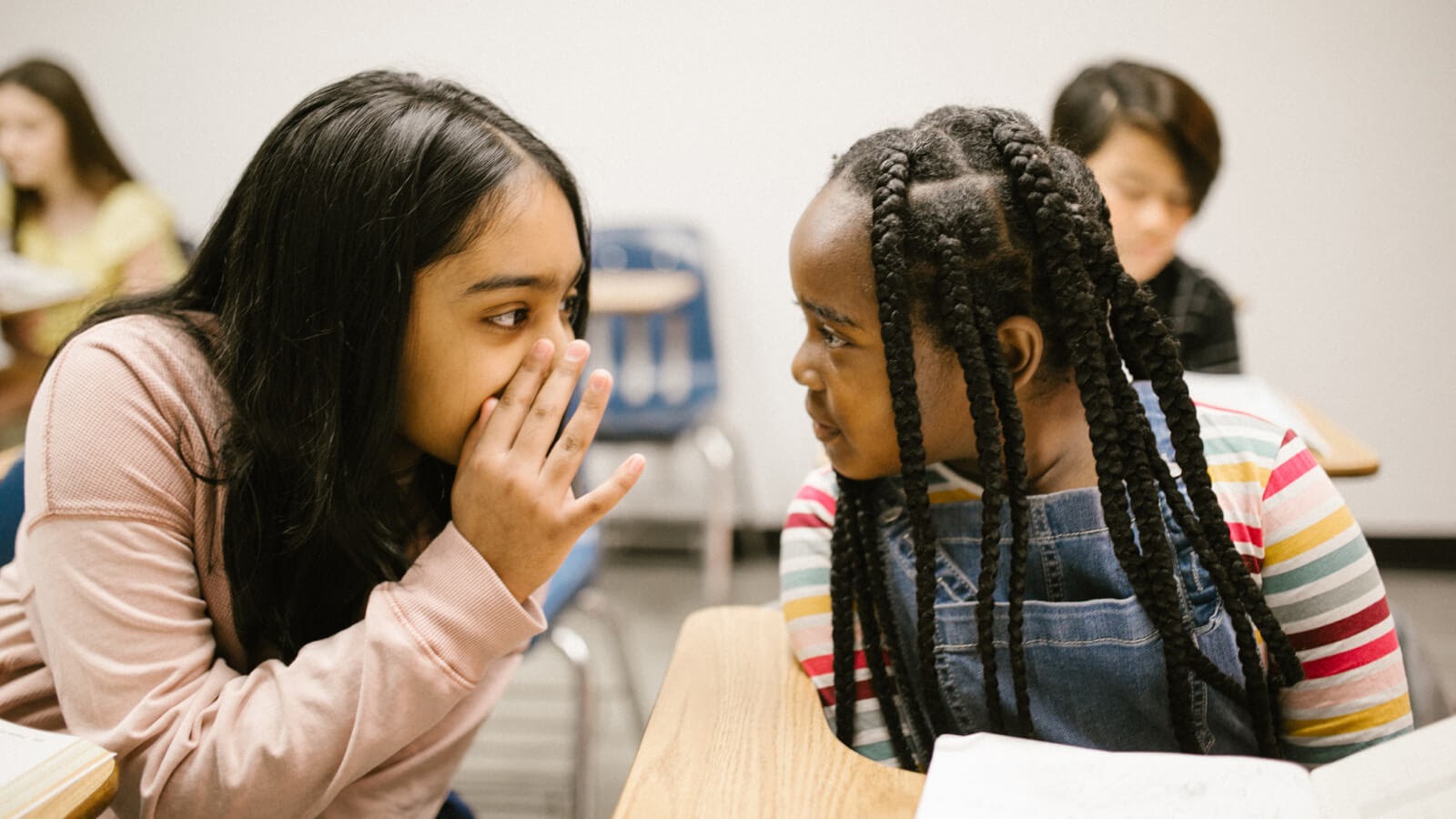 two school girls chatting in class sat at their desk, whispering to each other