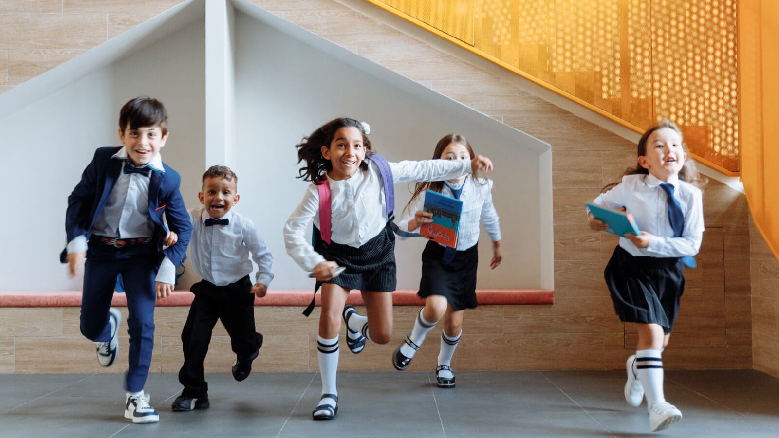 group of five school children running laughing in their grey and navy school uniform holding their school bags and books