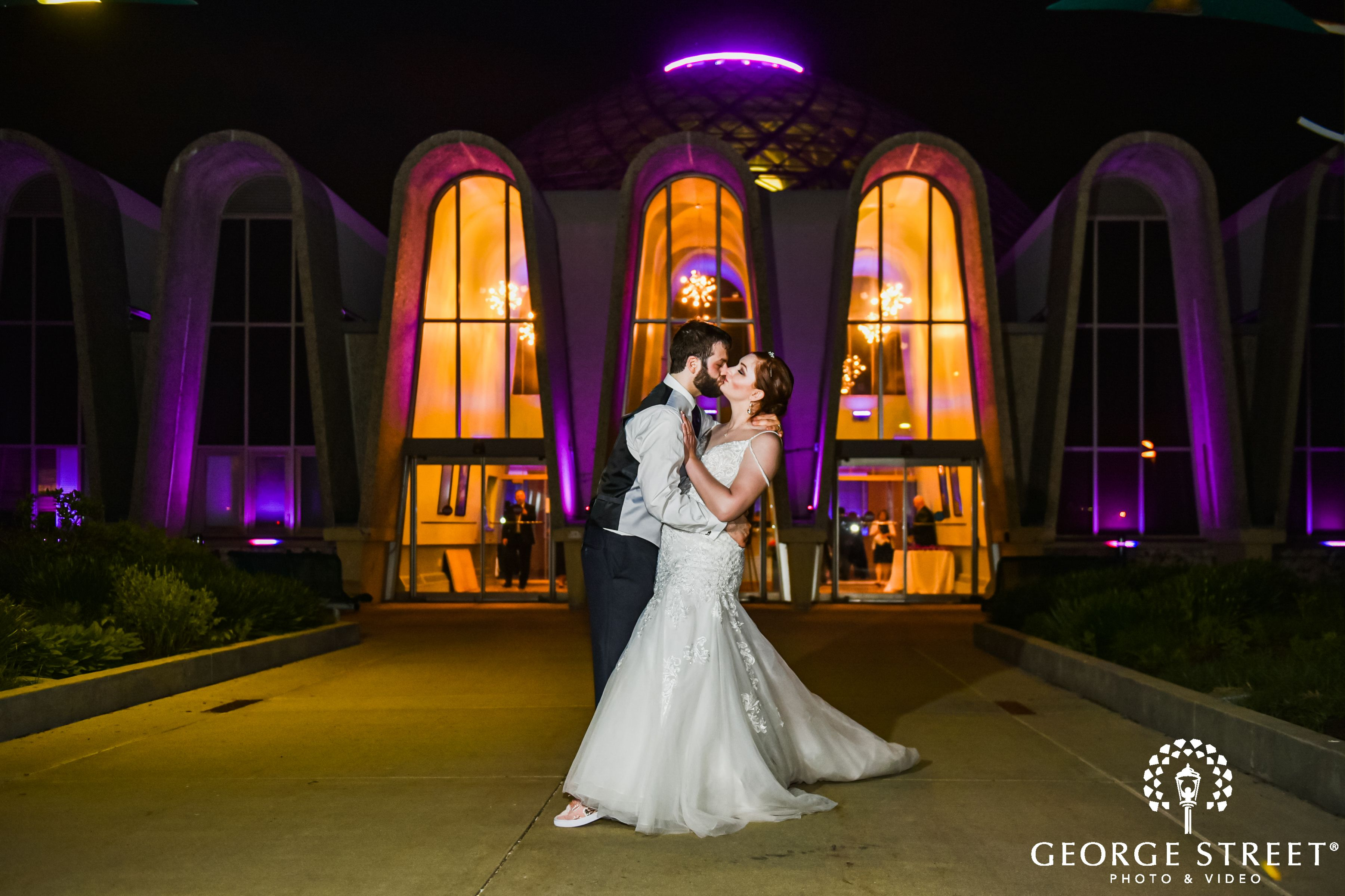 a man wearing formal wear kisses a woman in a wedding dress outside of the entrance to the Domes