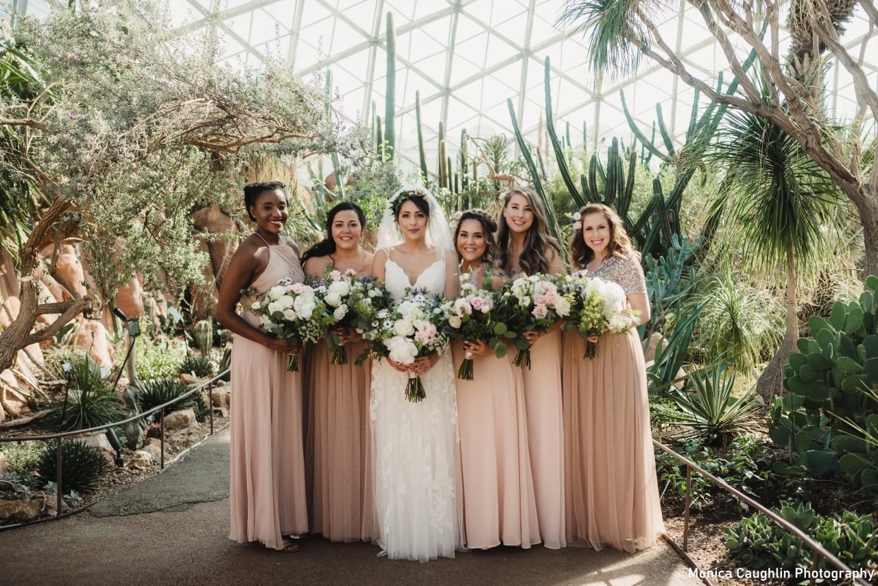 a group of five bridesmaids stand next to a bride in a wedding dress. All six women are holding bouquets and standing inside the desert dome