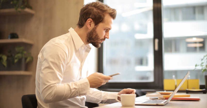 Man holding cell phone while looking at a laptop computer