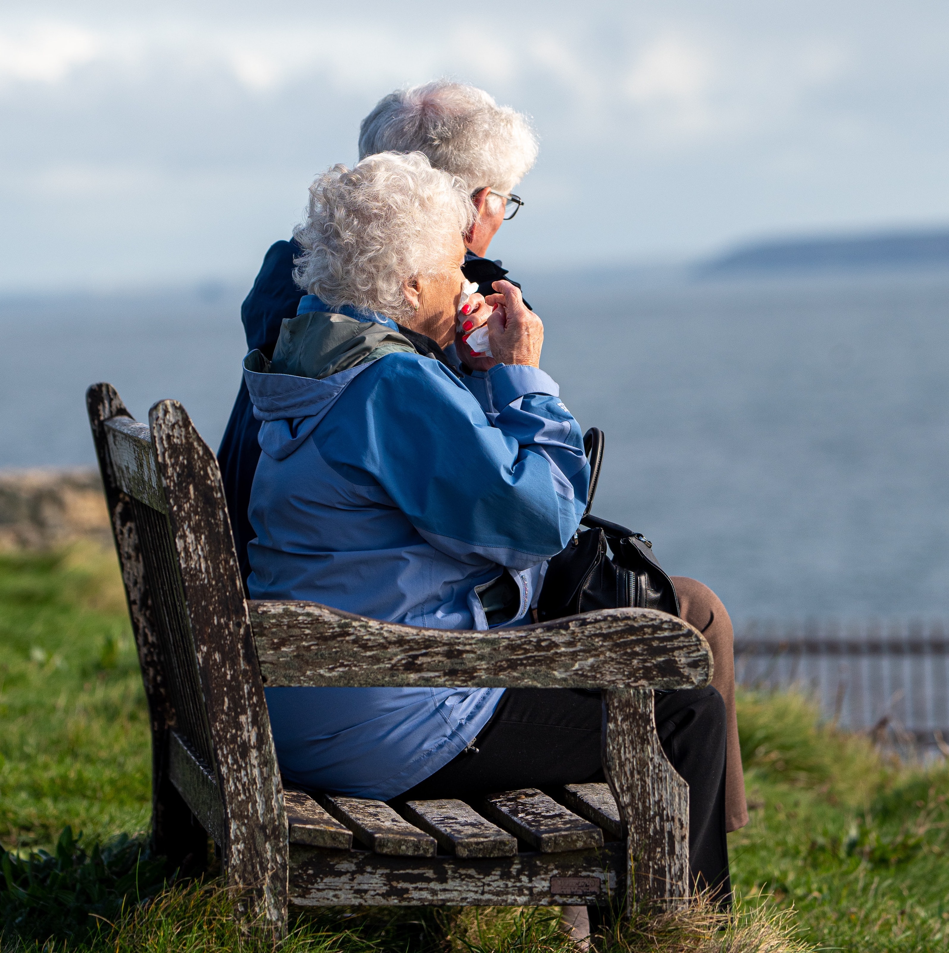 A elderly man and a woman sitting on a wooden bench by a large body of water.