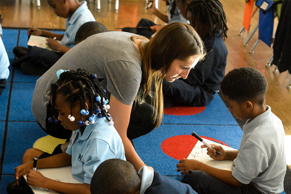 Teacher seated on floor with students, helping them with their work.