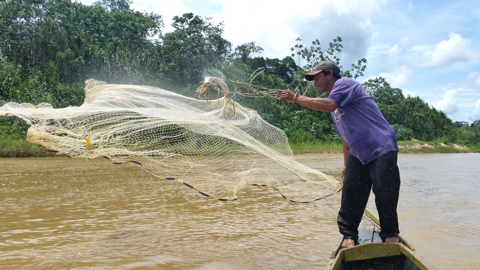 A man standing on a small boat throwing a fish net into the river