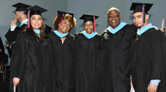 5 Hispanic and Black teachers smile at the camera with their arms around each other, wearing graduation gowns and hats. They have just received their masters of arts in teaching.