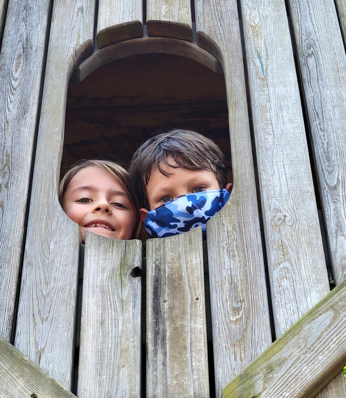 Two students peeking heads out of wooden jungle gym hole.