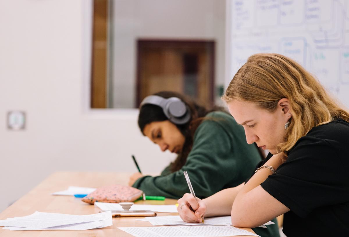 Two students focus on writing on papers strewn across their desk.