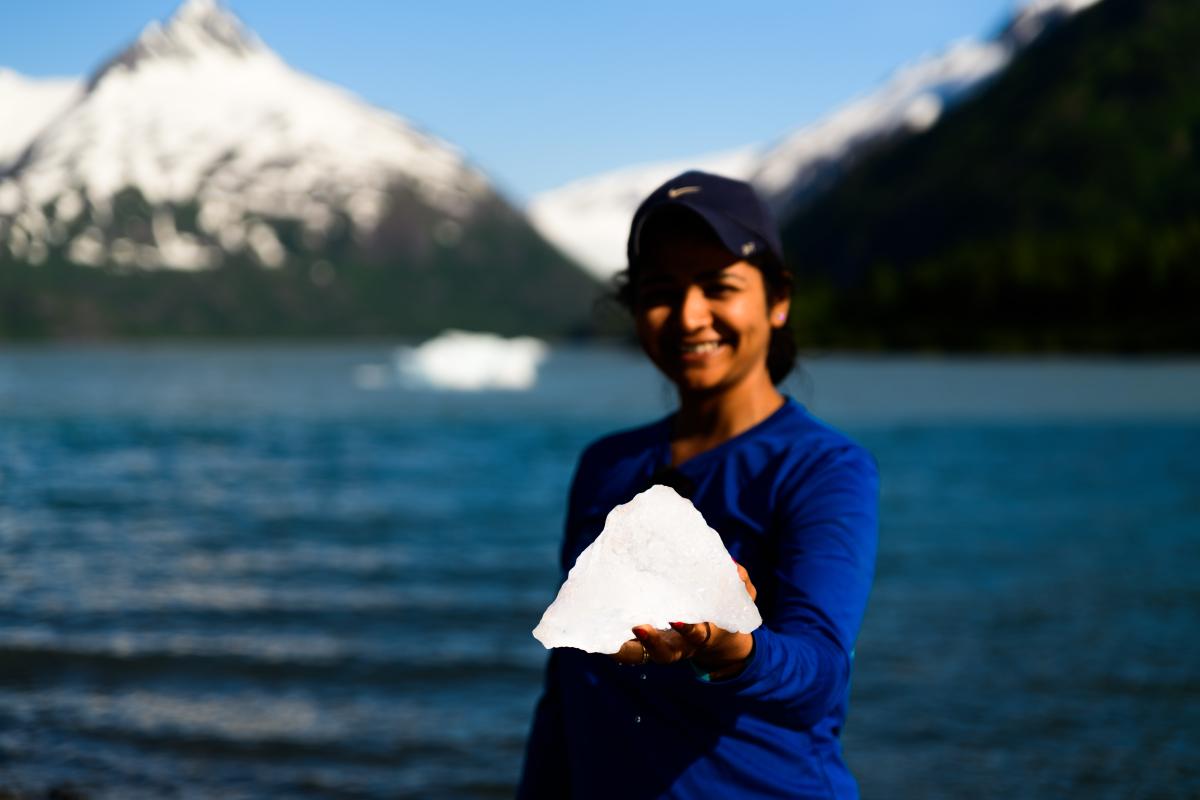 academic year in america student smiles and holds a handful of snow. Mountains and a lake are visible in the background.