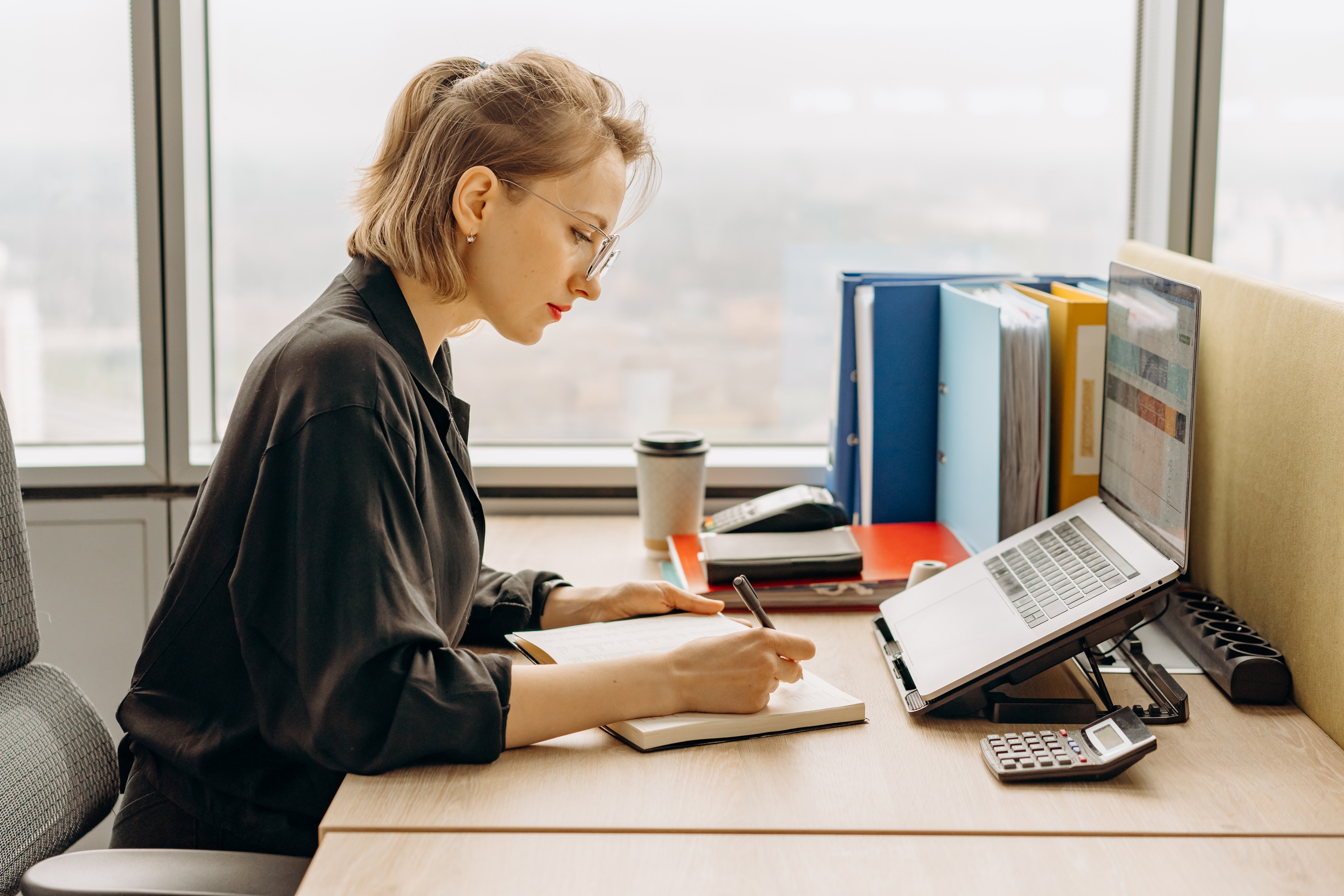 Woman in Black Long Sleeve Shirt Writing on a Notebook Near a Laptop