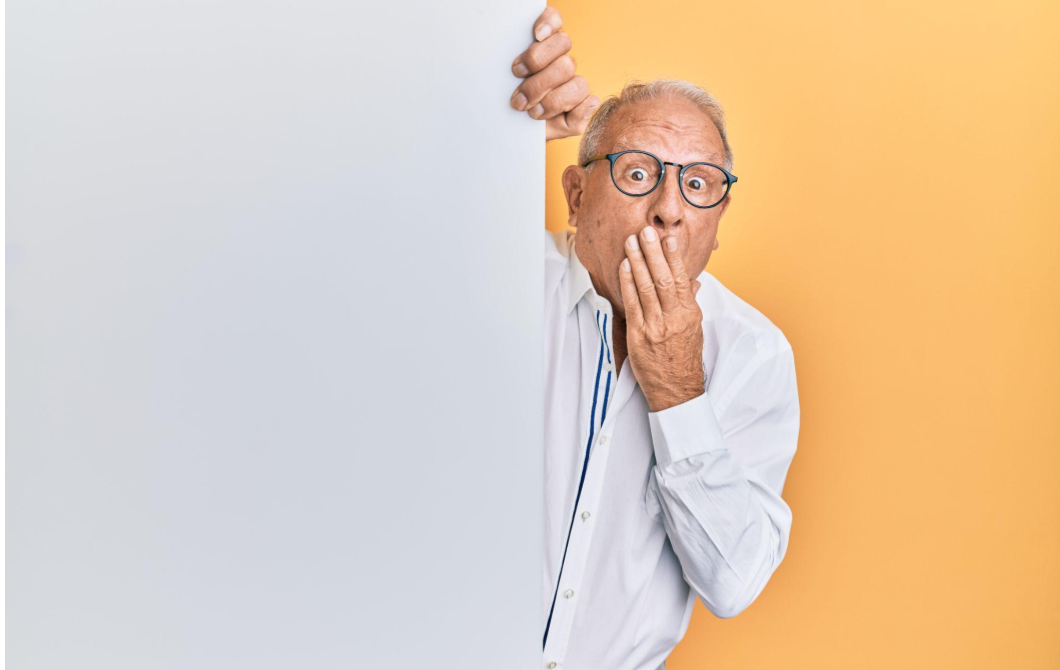 An older man peeks out from behind a wall, covering his mouth in fear