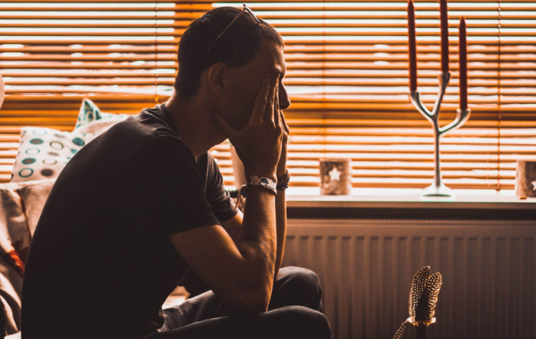 A depressed man sits half in darkness, hands over his face, thinking about therapy