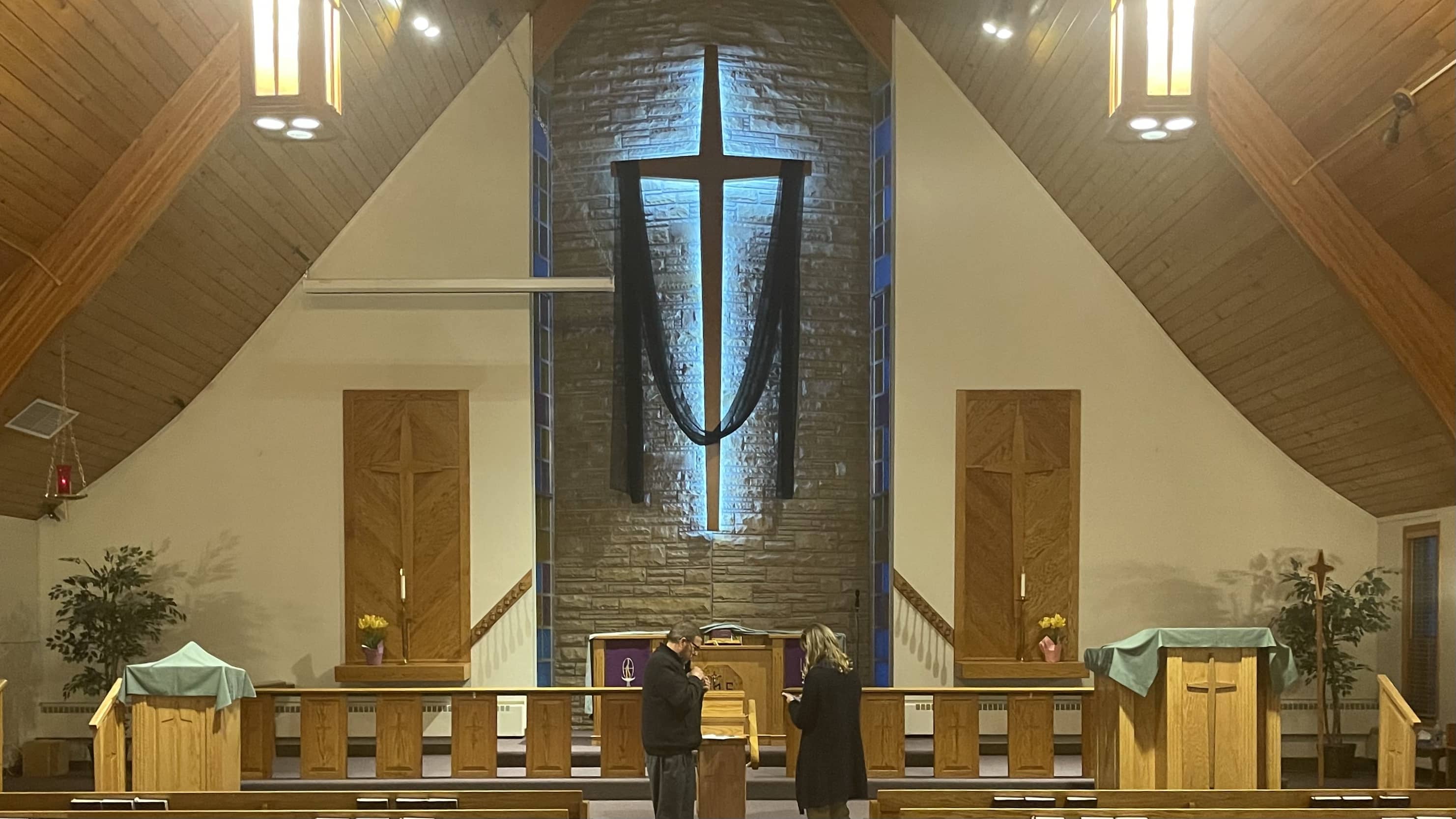 Image of a pulpit with a cross handing on the wall. The cross is backlit and hanging against brick