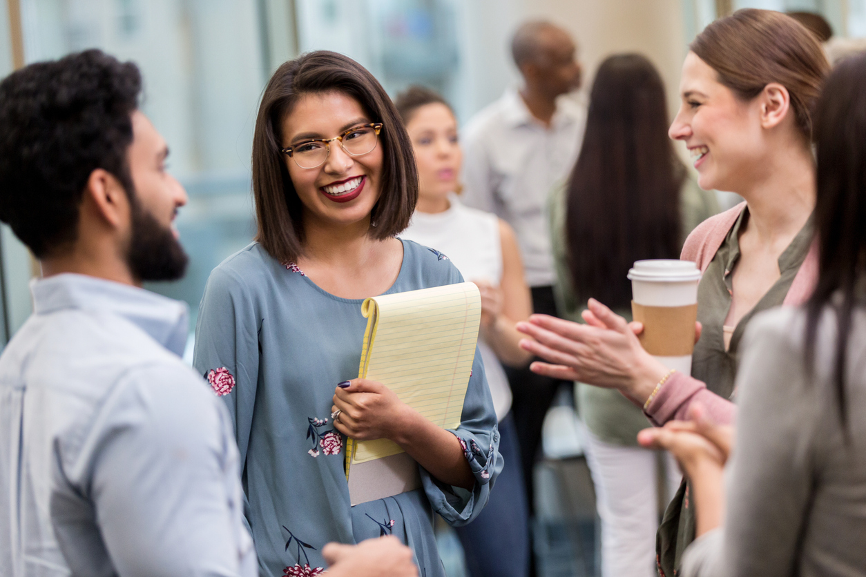 A young woman of south asian ethnicity holds a notepad and smiles at her colleague.
