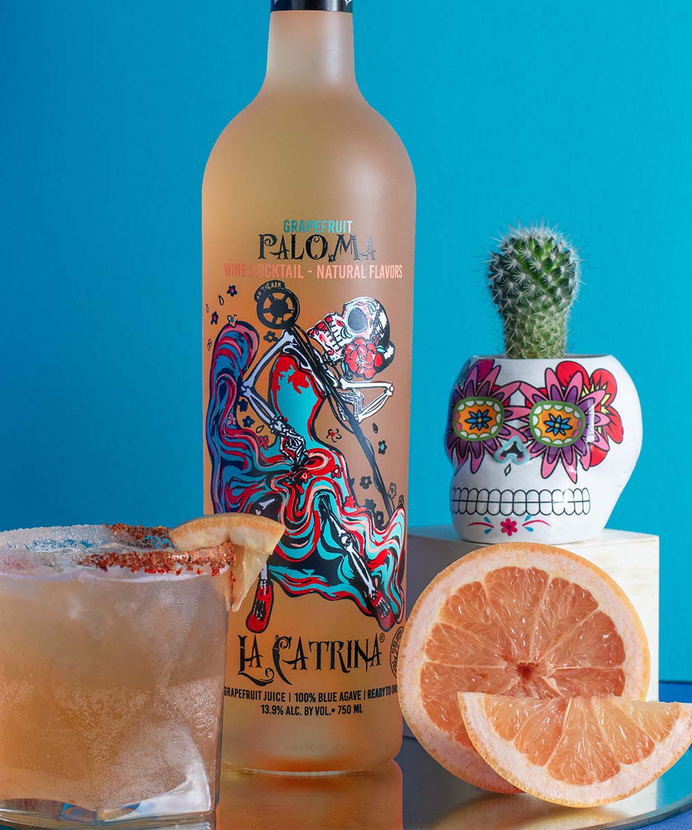 La Catrina Paloma Cocktail bottle next to a paloma glass, Day of the Dead skull and cut grapefruit