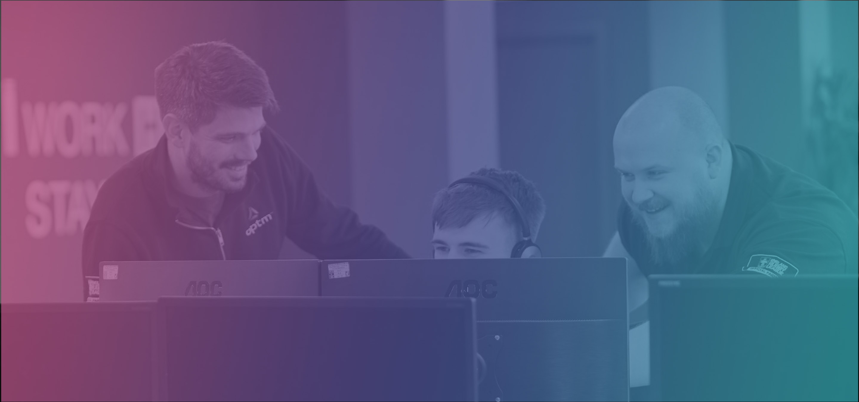3 men looking at the same screen with one sat down wearing headset and the others stood up next to him on either side