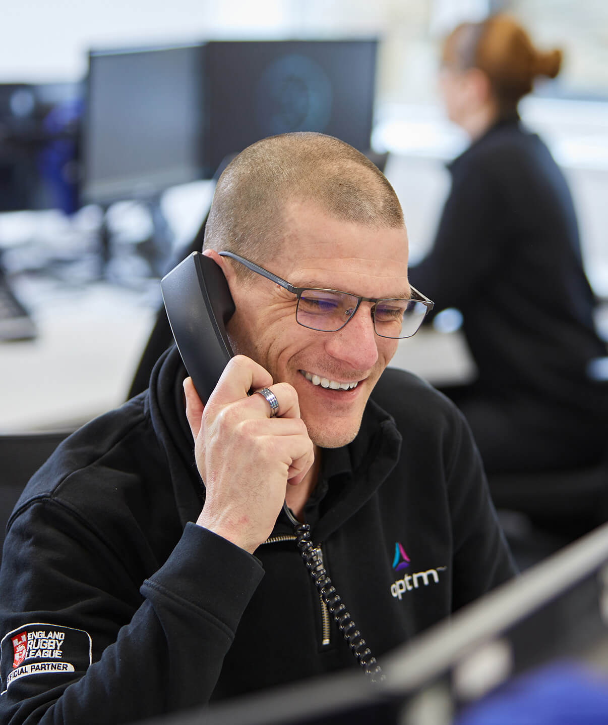 Man smiling on the phone wearing Optm jumper with woman in background out of focus