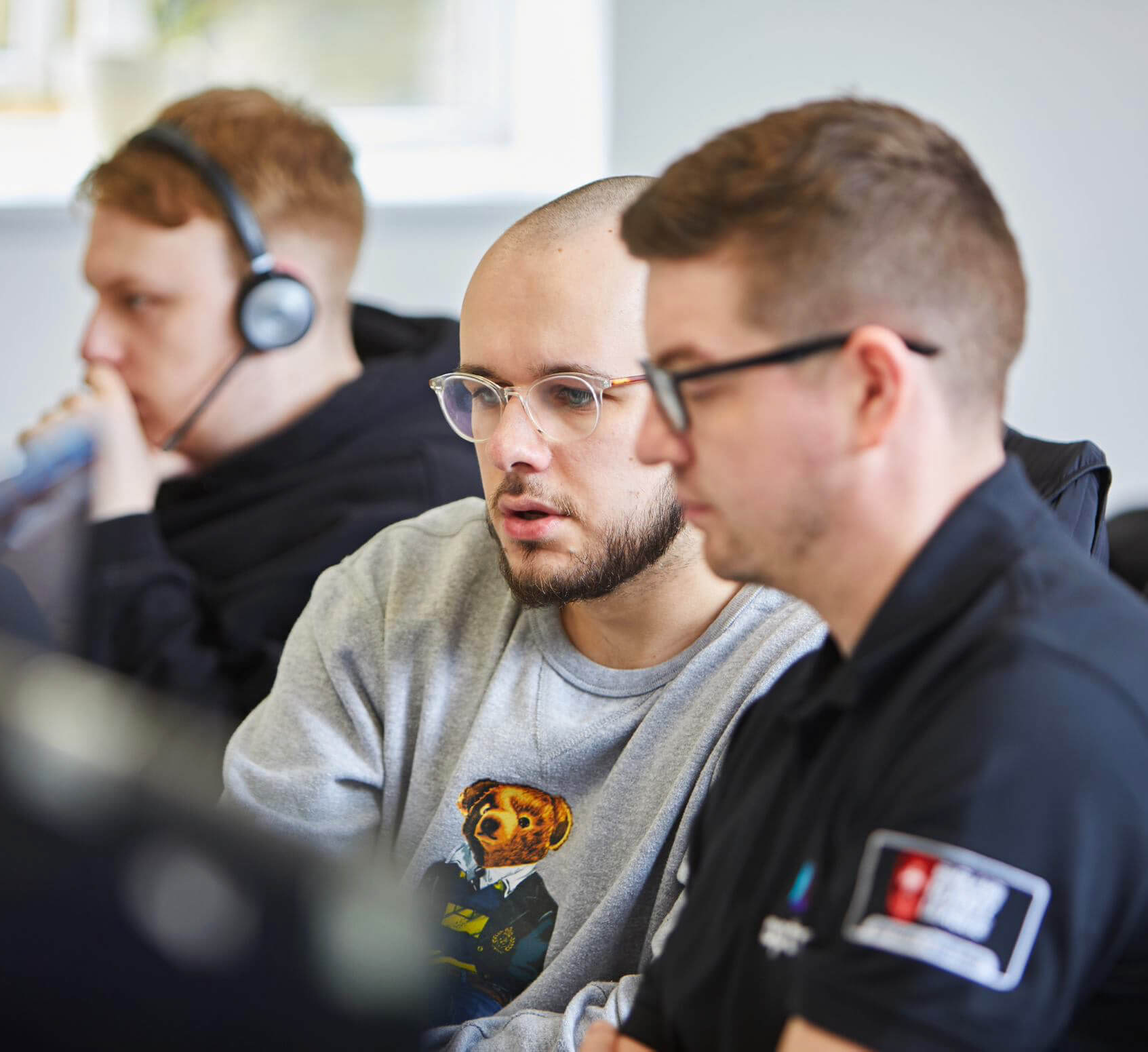 2 men sat next to each other looking in front at computer monitor with another person slightly out of focus wearing a headset
