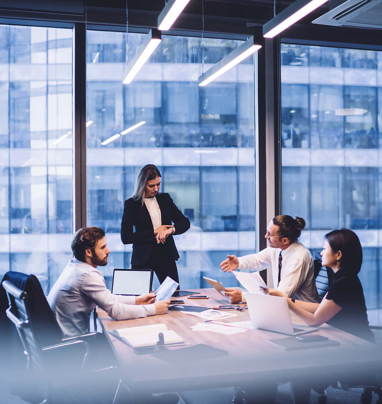 Three people sitting and one person standing at table in conference room with full height windows