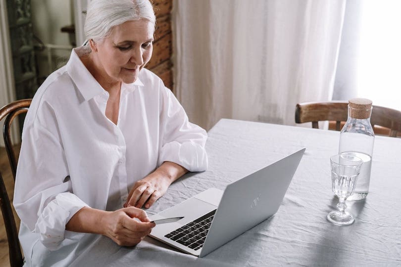 Seniorin in weißer Bluse am Laptop mit Patronus-Uhr beim Hausnotruf-Vergleich