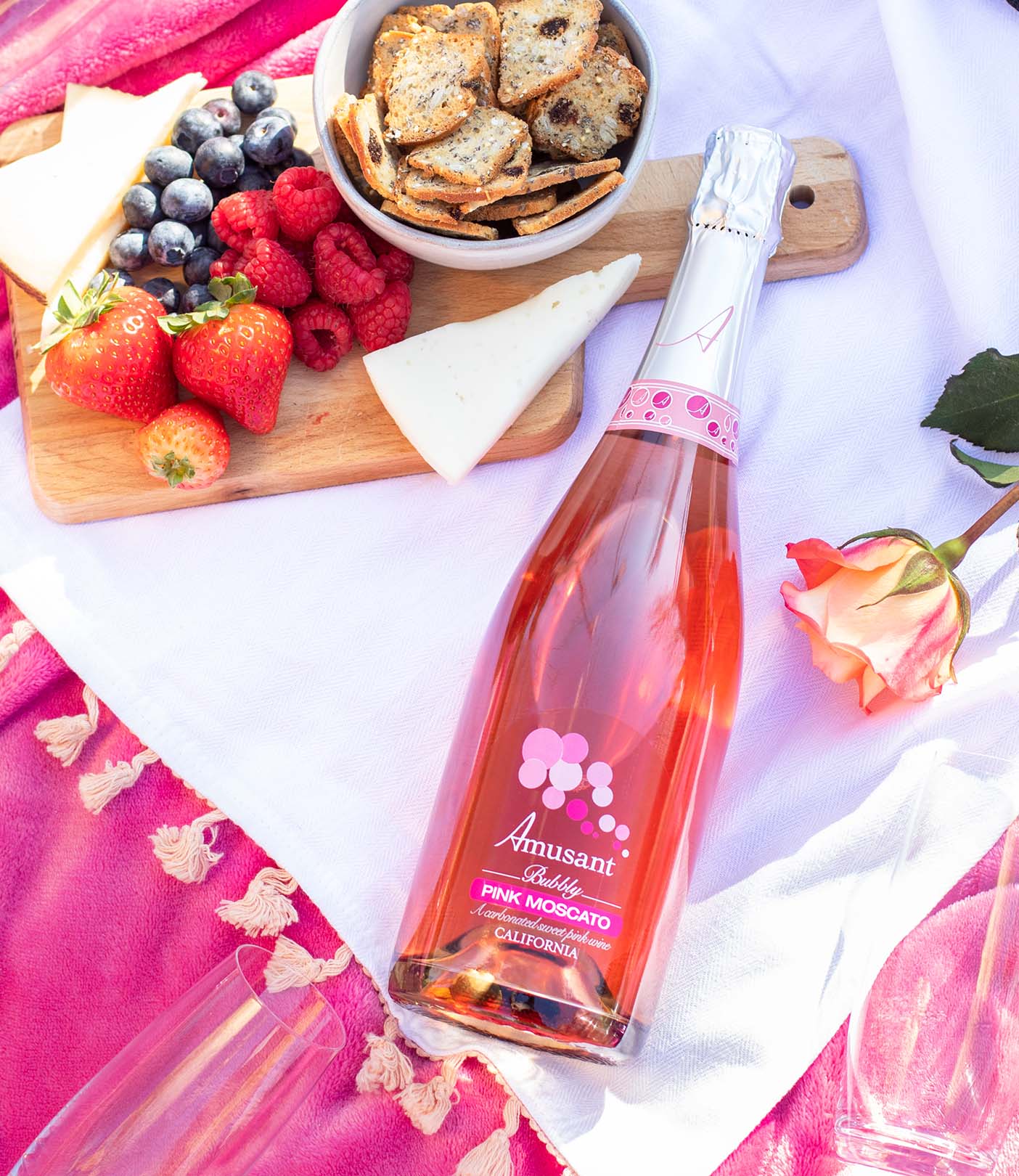 A bottle of Amusant Pink Moscato laying on a picnic blanket with fruit and snacks.