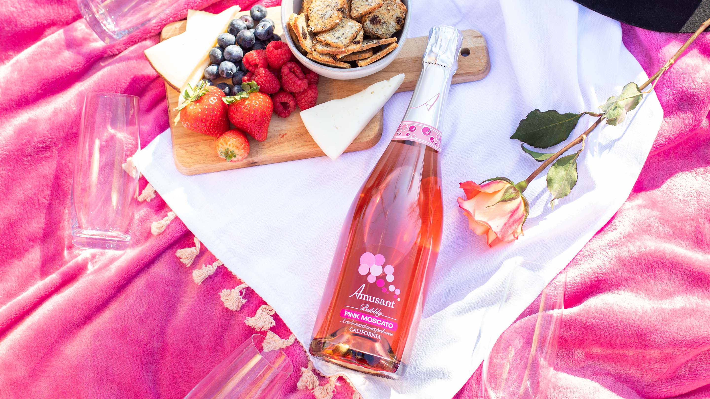 A bottle of Amusant Pink Moscato laying on a picnic blanket with fruit and snacks.