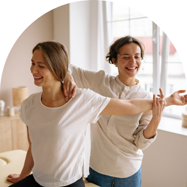 Two women smile as one helps the other stretch her arm and back.