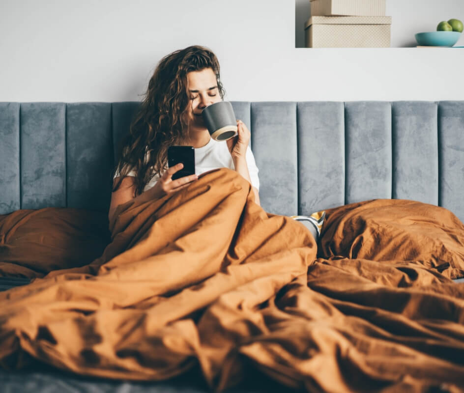 A woman smiles as she sips a beverage from a mug while relaxing in bed.