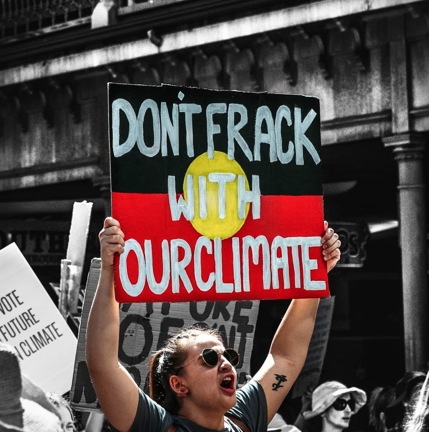 A black and white photo of Ebony at a rally, edited so that both Ebony and the sign she is holding appear in colour. Ebony is shouting and holding a sign that reads "Don't frack with our climate"