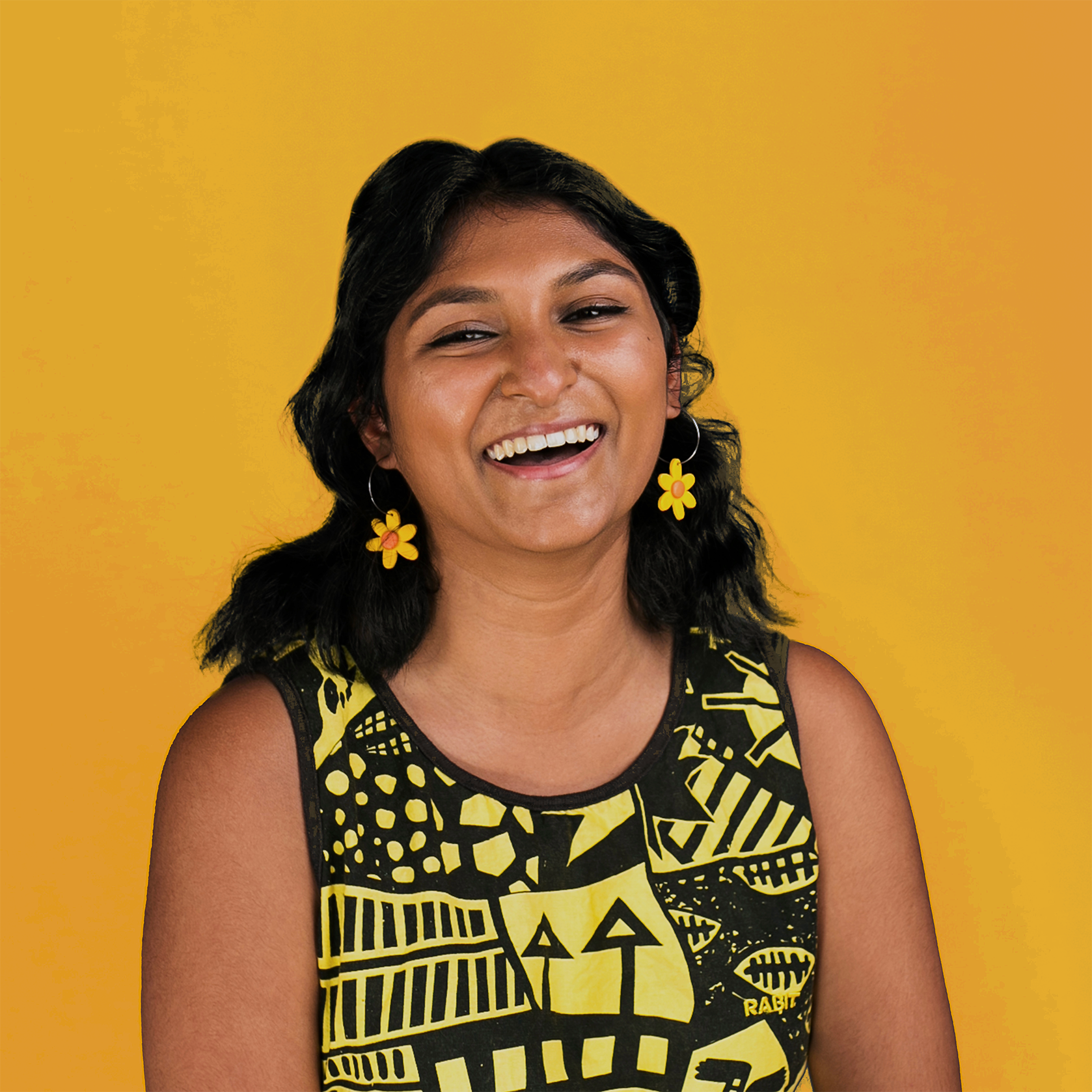 Grace is wearing yellow flower earrings and a yellow and black artwork shirt. She has shoulder length black hair and is laughing against a navy blue wall.