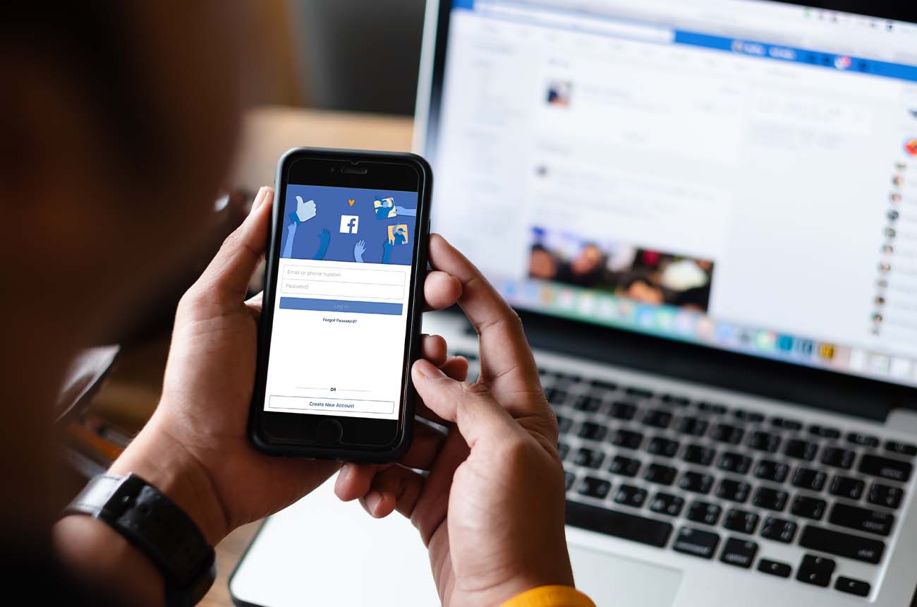 Man holding a cell phone logging into the Facebook app while looking at his laptop