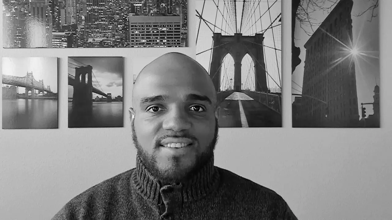 Black and white photo of André Givenchy sitting in front of a wall with photographs of famous bridges hanging on it.