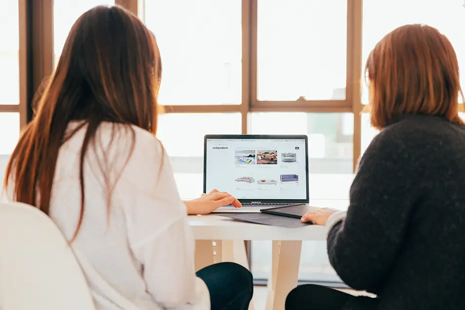 Two women sitting at a table online shopping on a MacBook Pro.