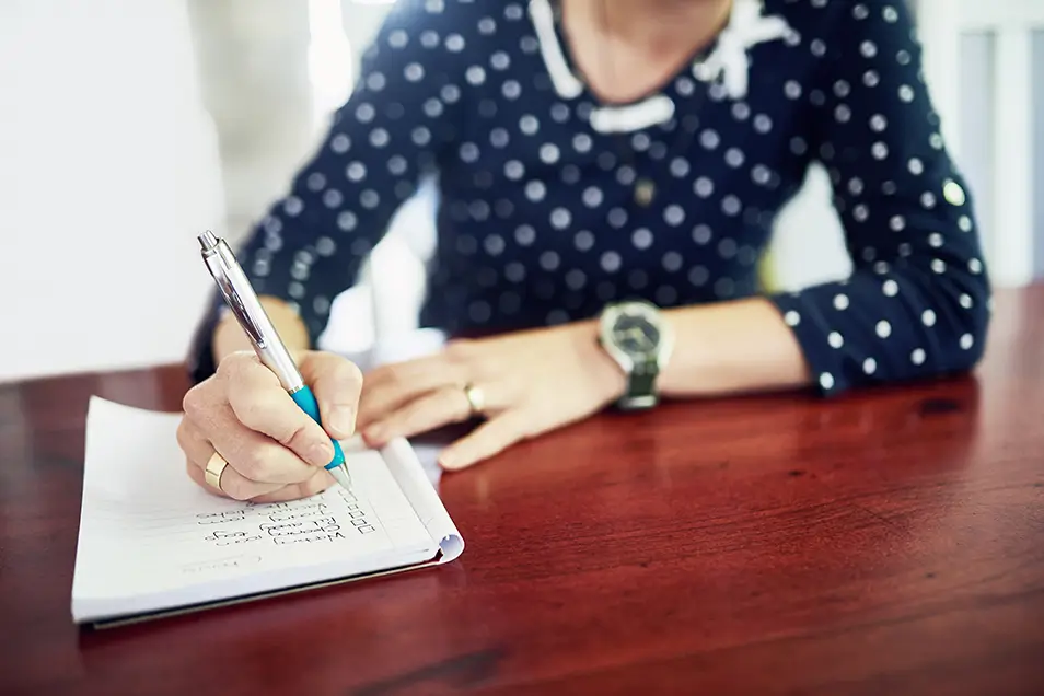 A woman sitting at a table making a to-do list on paper.