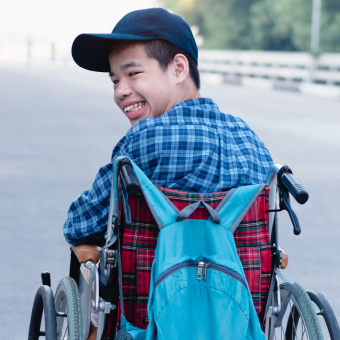 A boy in a wheelchair looking back at the camera wearing a plaid shirt and a baseball cap.