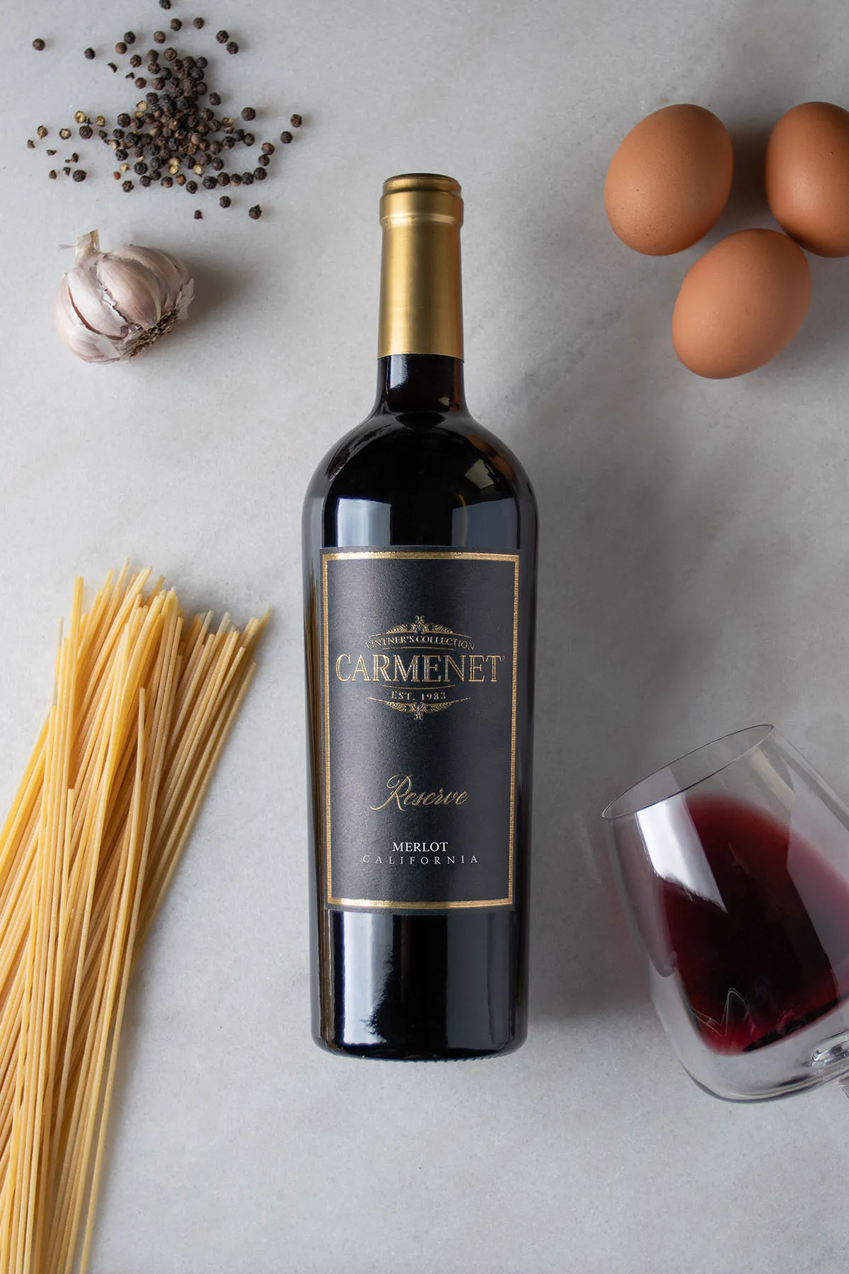 A bottle of Carmenet Merlot on a marble background with pasta, eggs, garlic and a tipped-over wine glass