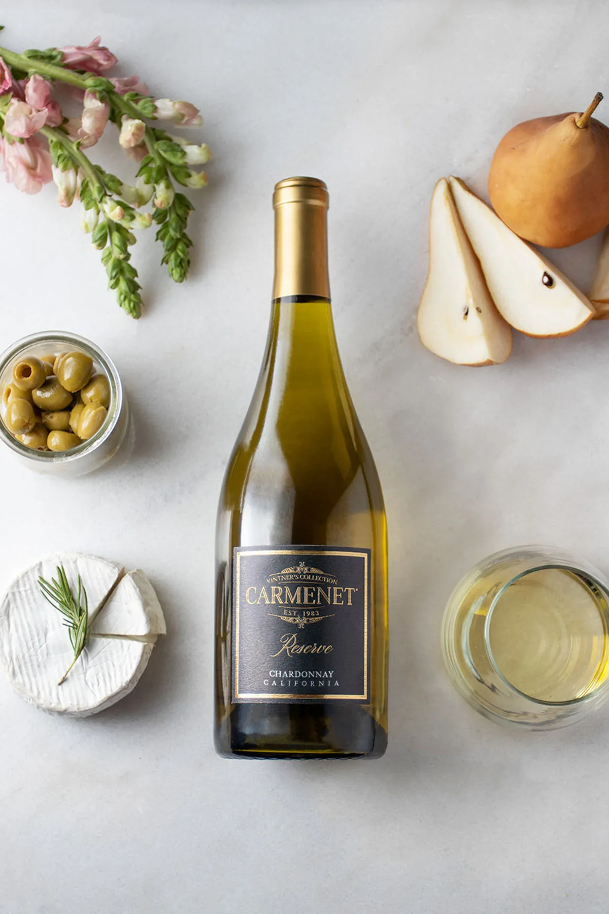 A bottle of Carmenet Chardonnay on a marble background with pears, nuts, cheese and a glass of wine
