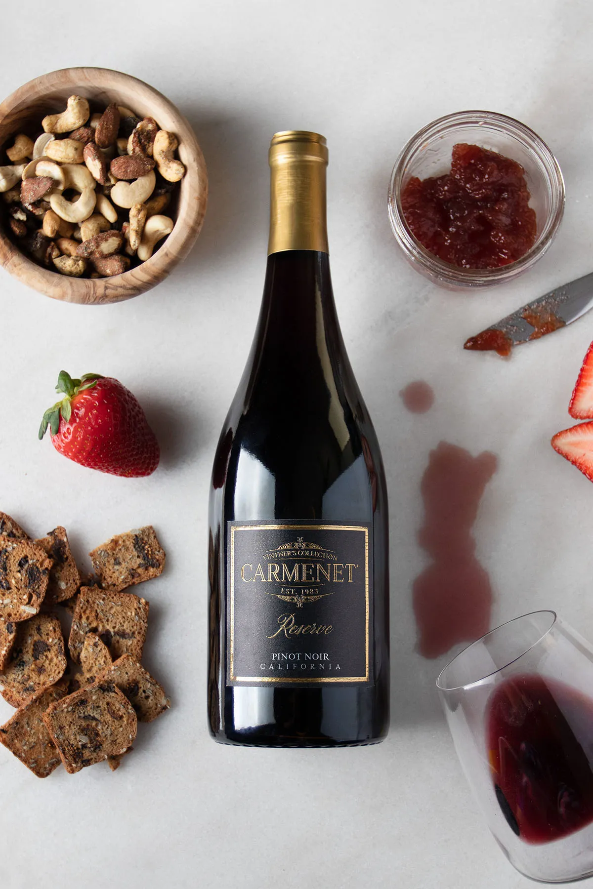 A bottle of Carmenet Pinot Noir on a marble background with fruit, nuts and a tipped-over wine glass