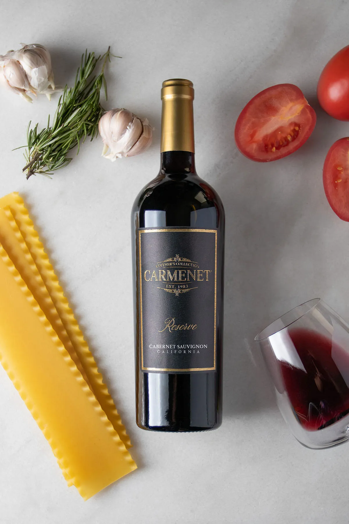 A bottle of Carmenet Cabernet Sauvignon on a marble background with pasta, fruit, garlic and a tipped-over glass