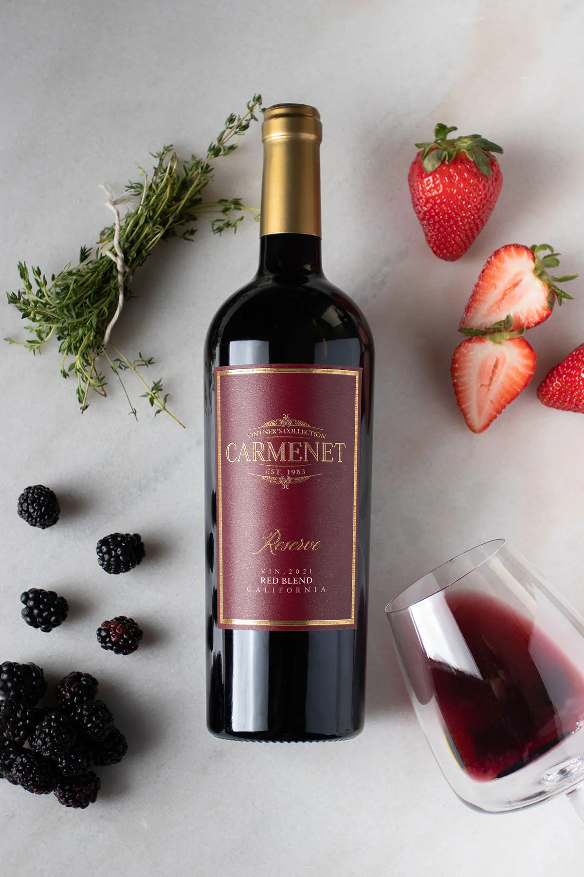 A bottle of Carmenet Red Blend on a marble background with fruit, herbs and a tipped-over wine glass