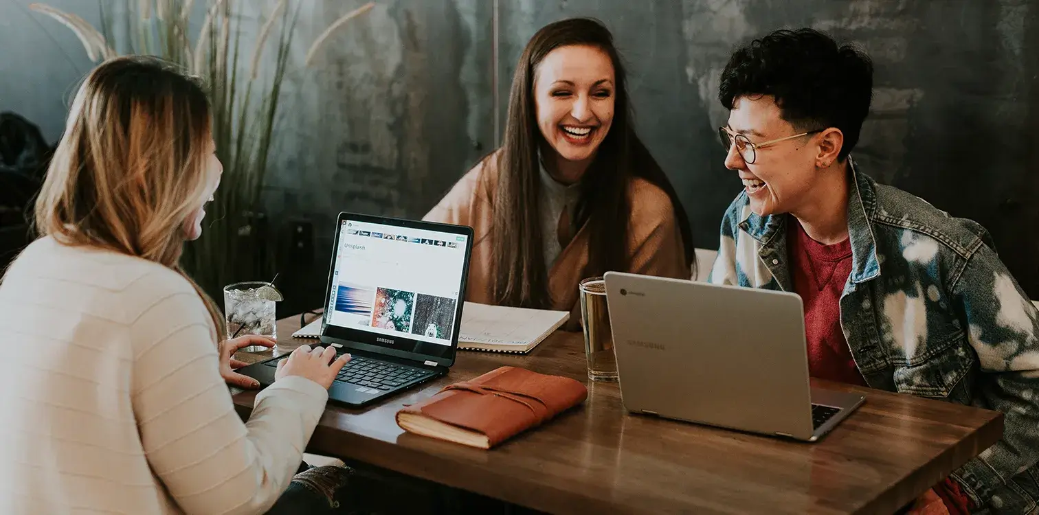 Drei Frauen sitzen mit Laptops an einem Konferenztisch und lachen.