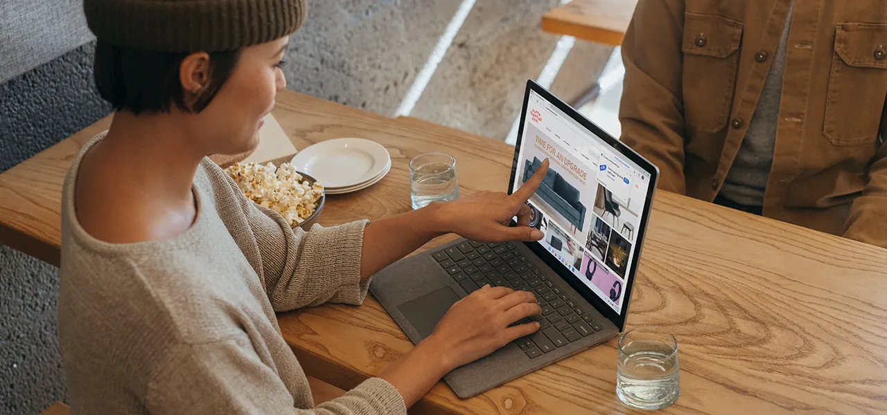 Frau im Pulli zeigt mit dem Finger auf ein Laptop Display am Esstisch.