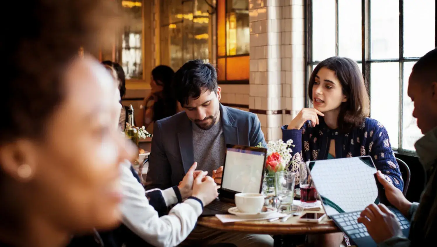 Mehrere Personen sitzen mit mobilen Geräten in einem Café am Tisch und unterhalten sich.