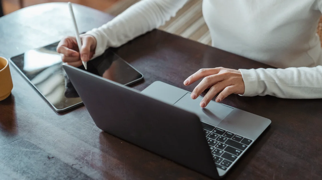 Person am Tisch, die am Laptop sitzt und daneben ein Tablet bedient