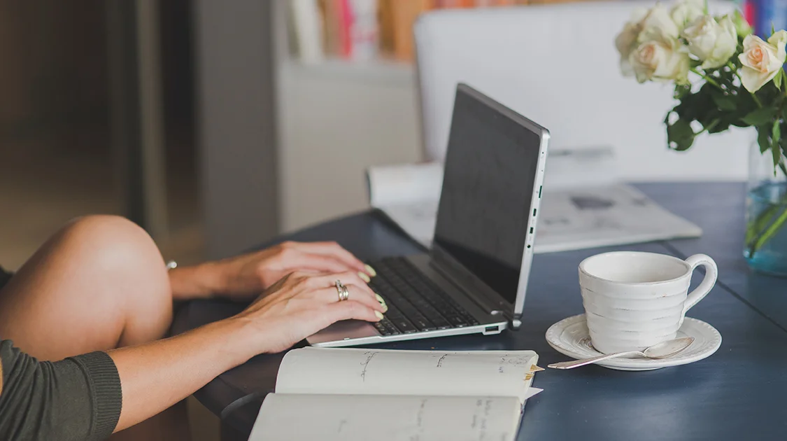 Person mit Laptop und Kaffee an einem Tisch im Homeoffice und das Knie angewinkelt