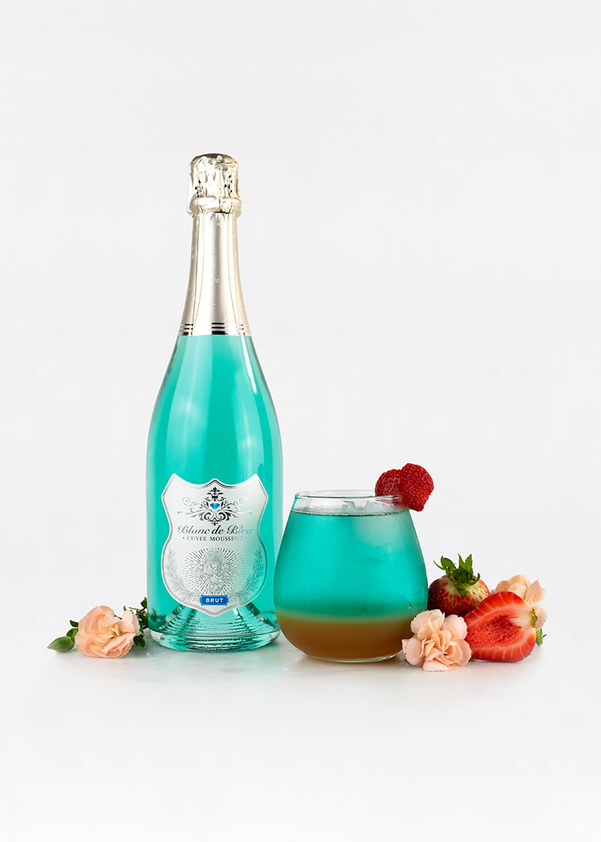 A "Pinky Out" cocktail next to a bottle of Blanc de Bleu Brut wine, with strawberries and flowers around the drink and bottle.