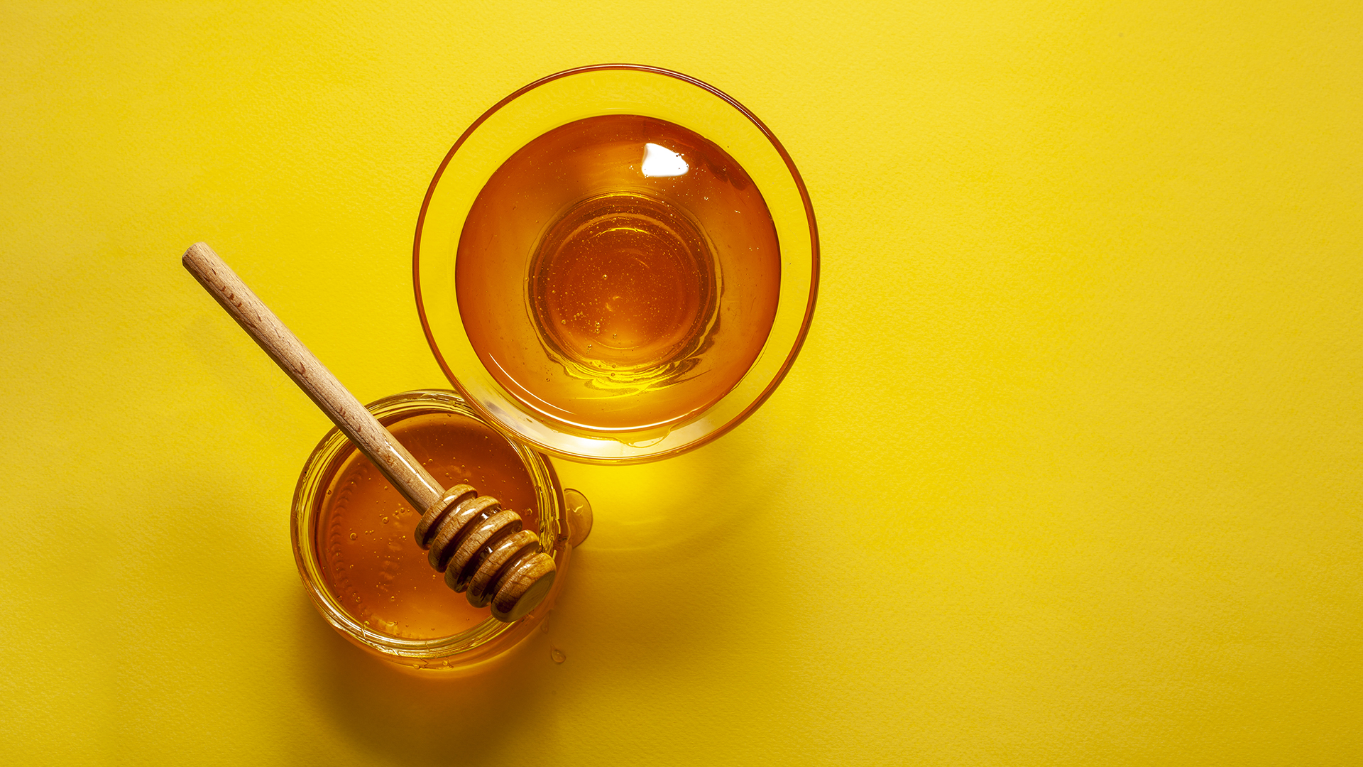 A imagem com fundo amarelo apresenta dois potes de tamanhos diferentes contendo mel. Um deles, o menor, possui um pegador de mel em cima escorrendo uma pequena quantidade de mel.