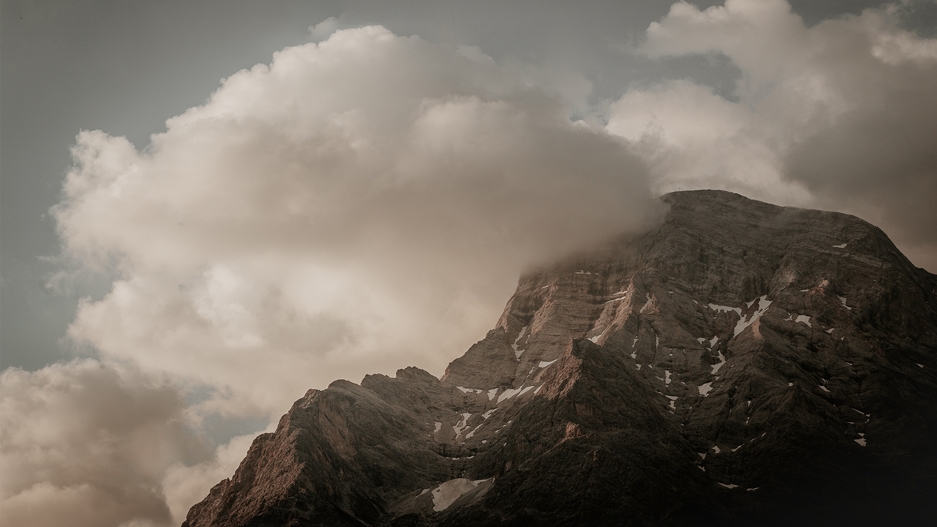 A imagem apresenta uma fotografia na qual nos é apresentado o cume de uma montanha em tons de cinza e marrom, com densas nuvens cobrindo parcialmente a parte superior da montanha. O céu, ao fundo, também é cinza.