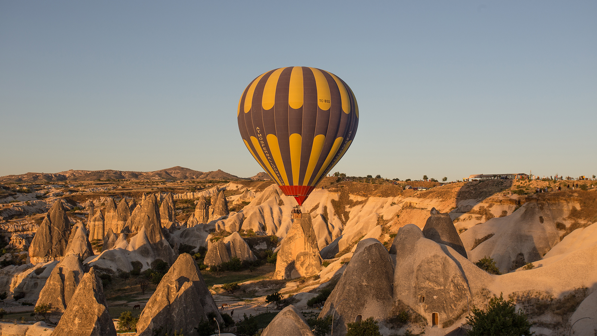 A imagem apresenta uma fotografia colorida de um balão de ar quente nas cores amarelo e azul, no meio de grandes rochas circundantes. O céu está azul e sem nuvens e a folhagem terrestre rasa e seca, na cor verde claro.