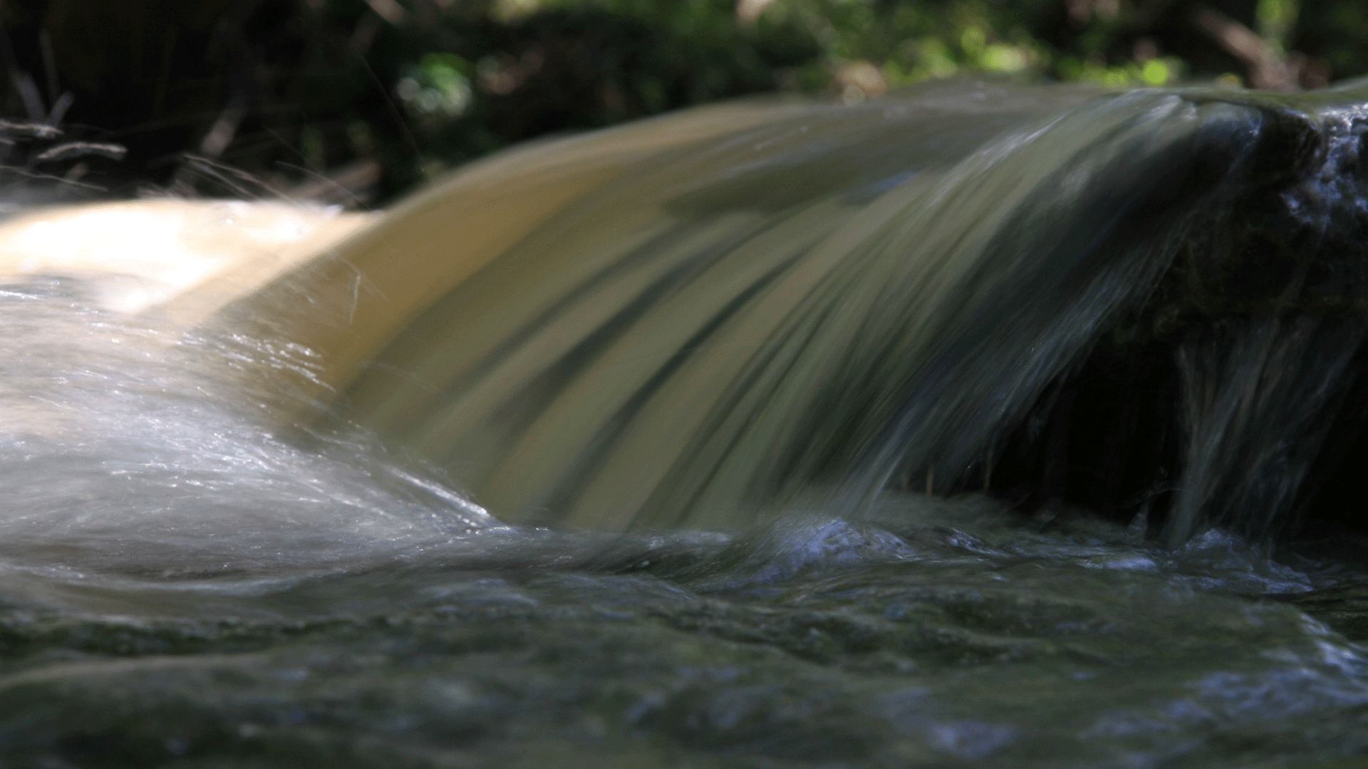 A imagem apresenta uma fotografia, nela, vemos uma corrente de água cristalina advinda de um rio em uma pequena queda. No fundo, enxergamos folhagens borradas.